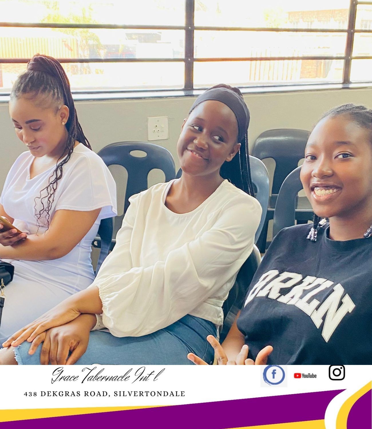 Three young women seated indoors, two smiling at the camera, one looking at her phone.